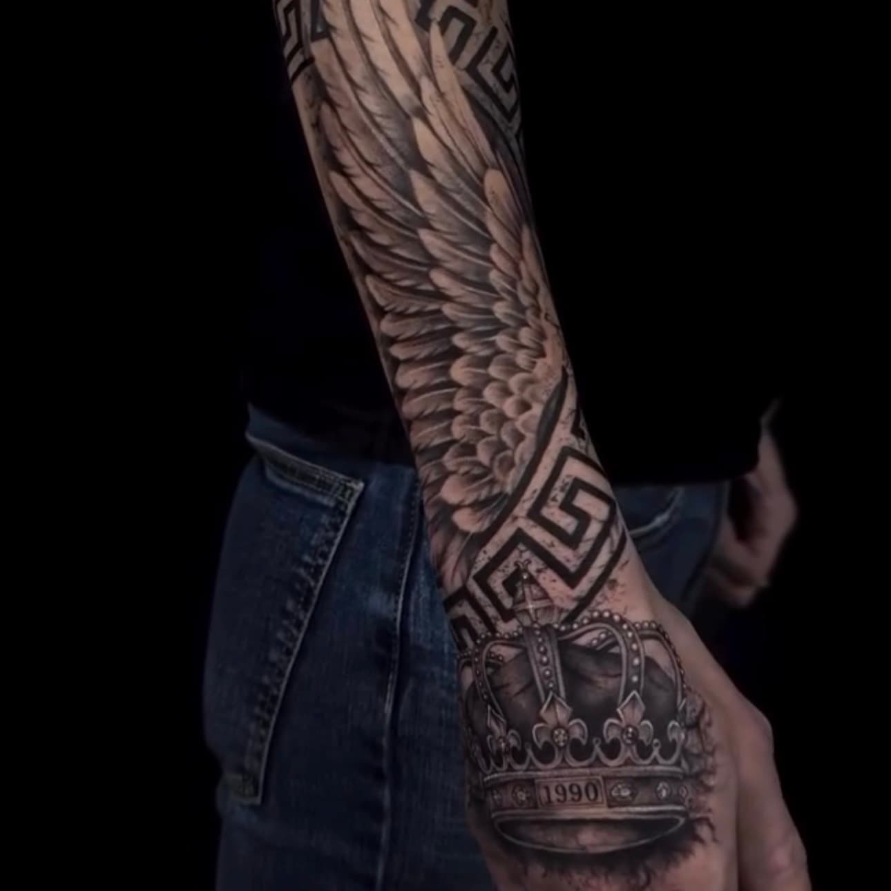 Tatuaje de alas realistas con greca y corona en la mano